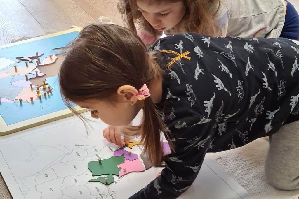 Două fetițe pe podea asamblând harta puzzle din lemn a continentelor — activitate de geografie la grupa 3-6 ani